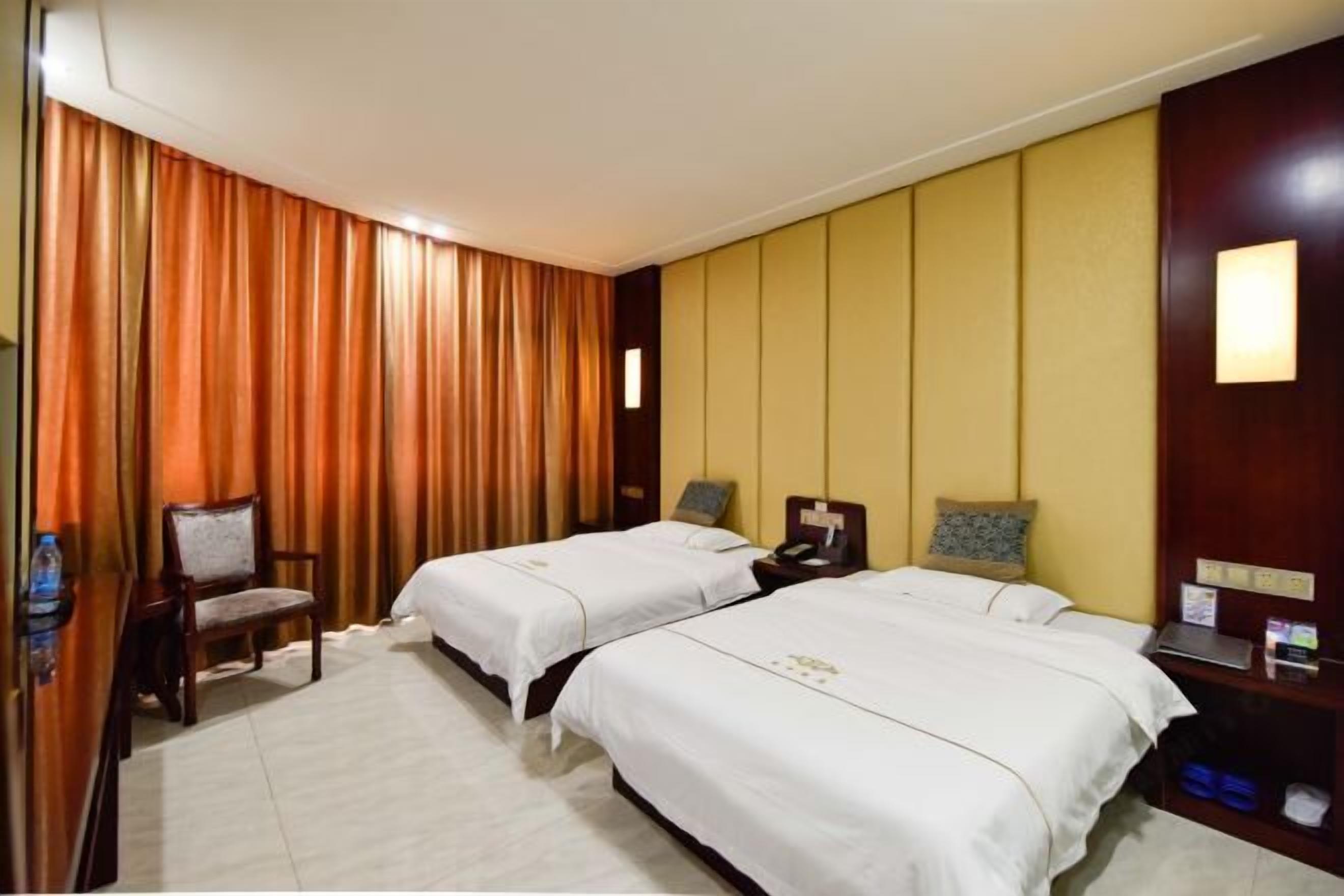 A hotel room with two beds, a wooden chair, a nightstand with a lamp, and a wall-mounted shelf.