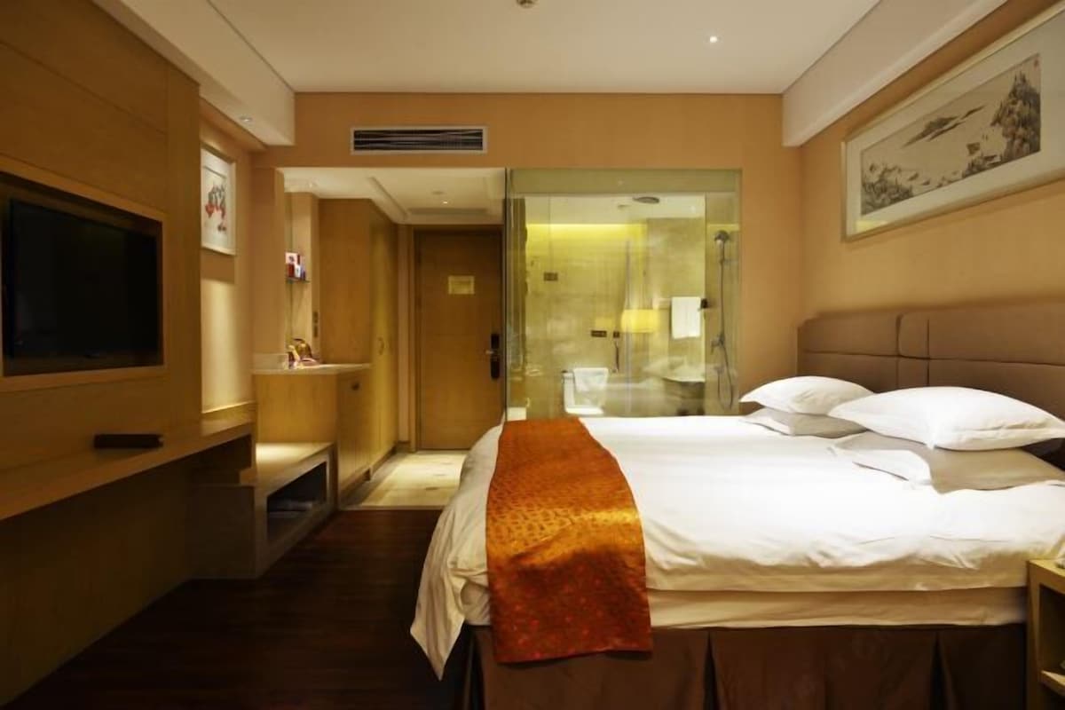 A hotel room with a bed, a television, a bathroom with a glass partition, and a painting on the wall.
