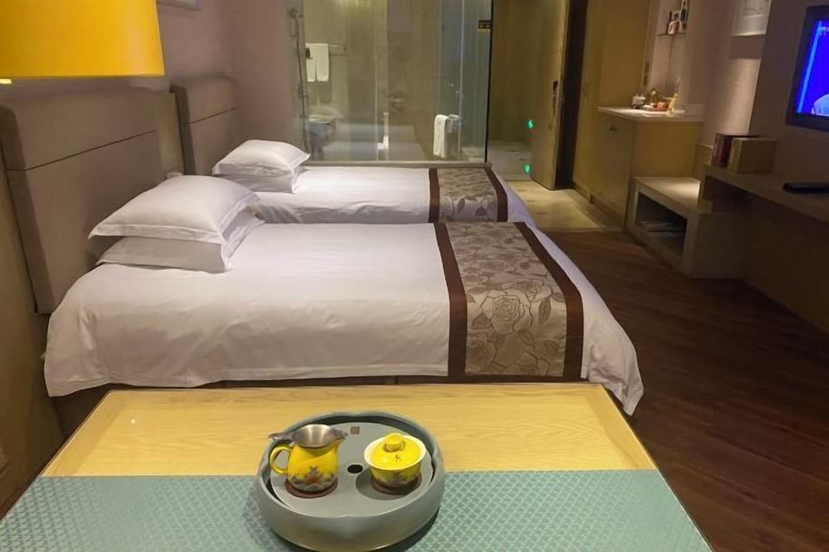 A hotel room with two beds, a shower area, a small table with a teapot and cup, and a TV on the wall.