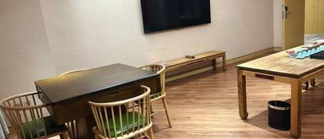 A room with a wooden table, two chairs, a bench, a television on the wall, and a trash can.