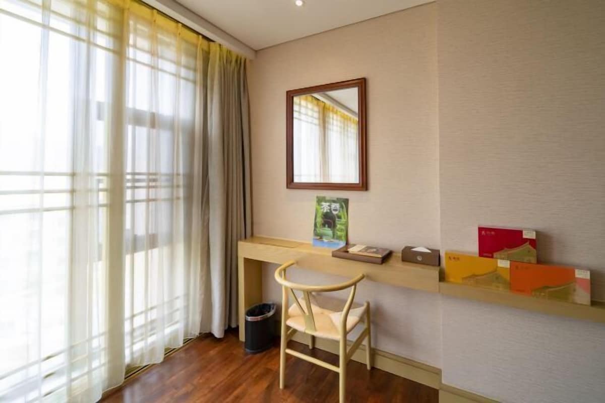 A hotel room with a wooden desk, a chair, a mirror, and a shelf with books and a small trash bin.