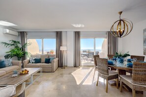 A modern living room with a sofa, coffee table, dining area with wicker chairs, and a view of the ocean through large windows.