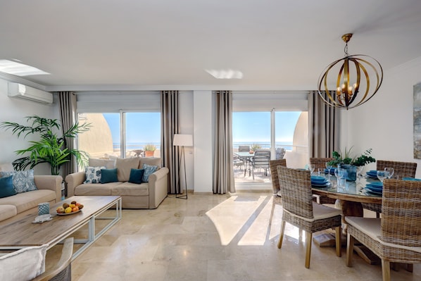 A modern living room with a sofa, coffee table, dining area with wicker chairs, and a view of the ocean through large windows.