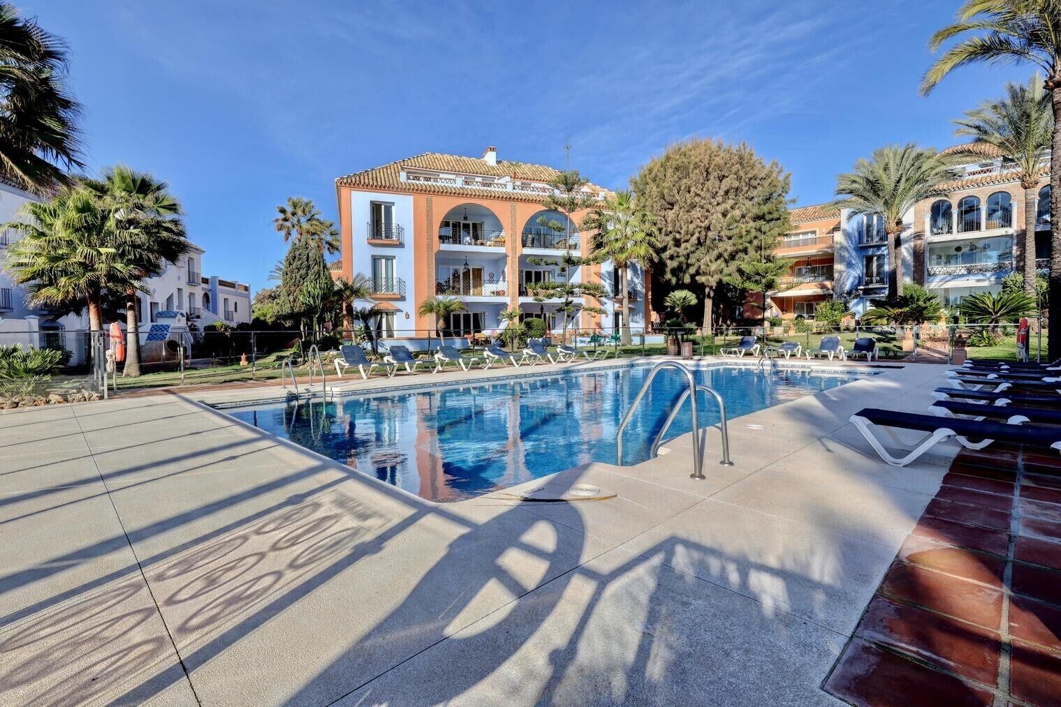 Une piscine avec des chaises longues, des palmiers et un bâtiment doté de fenêtres en arc de cercle.