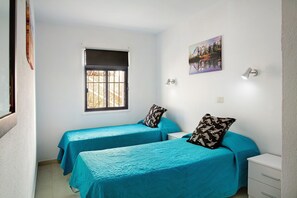 A bedroom with two beds, a window, a painting, and a bedside table.