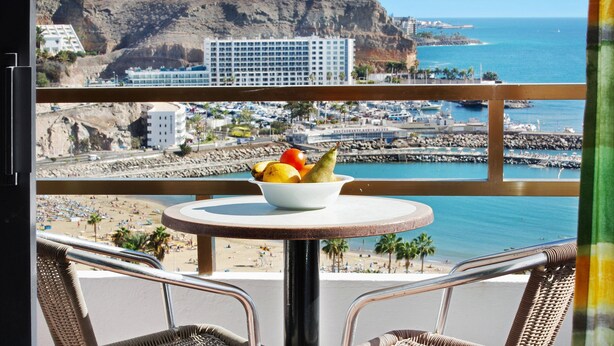 A balcony with a table holding a bowl of fruit, overlooking a beach, a marina, and a hotel. - Mirra Apartment – Beach View (Puerto Rico)