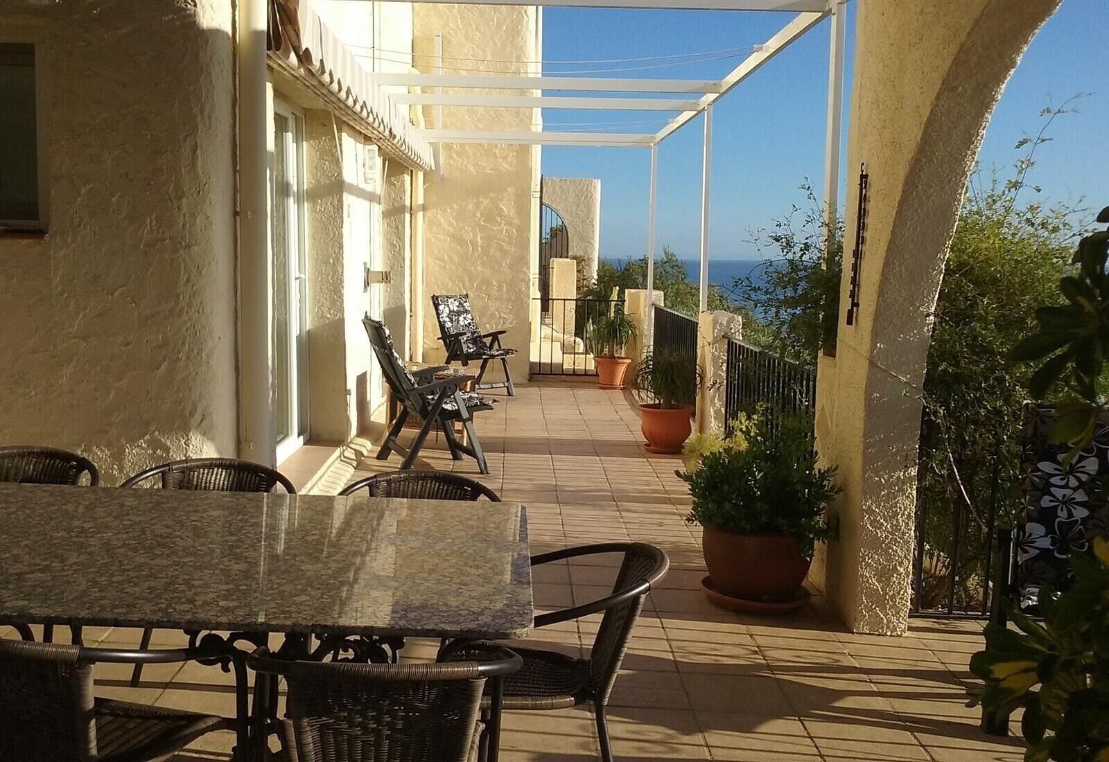 An outdoor patio with a table and chairs, potted plants, and a view of the sea.