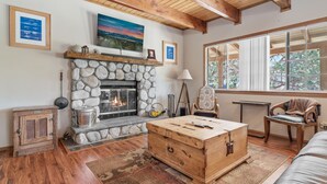 A cozy living room with a stone fireplace, wooden furniture, and a large window offering a view of the outdoors. - Hikers Haven - Close to Hiking, Fireplace, Horseshoes, Deck, BBQ (Big Bear City)