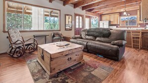 A spacious living room with a wooden coffee table, leather sofa, and rocking chair. A large window provides natural light, and a kitchen area is visible in the background. - Hikers Haven - Close to Hiking, Fireplace, Horseshoes, Deck, BBQ (Big Bear City)