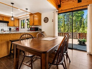 A wooden dining table with chairs, a kitchen with wooden cabinets, a red microwave, and a view of trees through a sliding glass door.