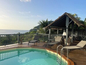 Ein Poolbereich mit Holzterrasse, Liegestühlen, einer kleinen Laube und Blick auf den Ozean.