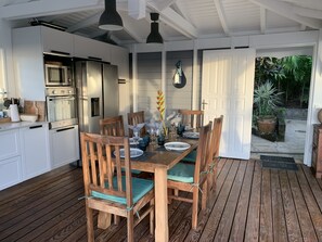A modern kitchen with white cabinets and stainless steel appliances, a wooden dining table with chairs, and a view of a garden through an open door. - Beautiful Villa with Sea View & Pool - Relaxing stay in the tropics (Deshaies)