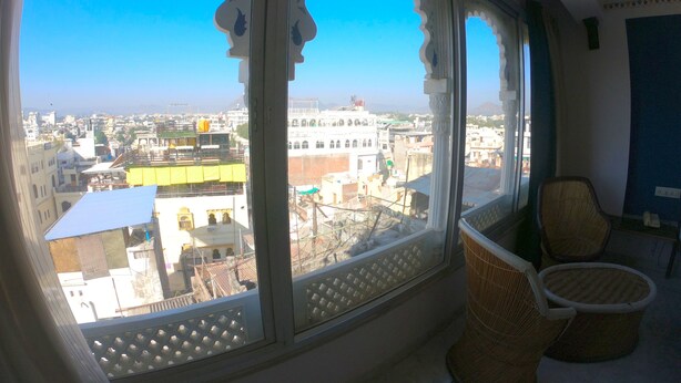 A room with a large arched window offering a city view. There are wicker chairs and a small table. - Little Peepal House (Udaipur)