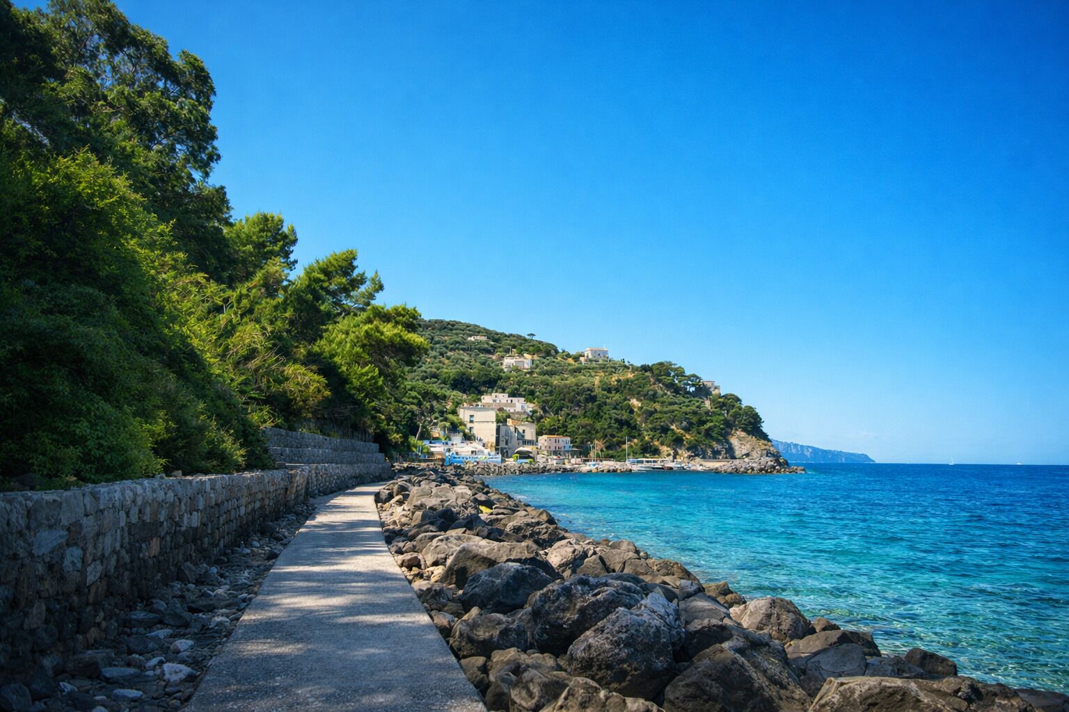 Un camí costaner amb una paret de pedra a un costat i un litoral rocos a l'altre, que condueix a una vessant amb edificis.