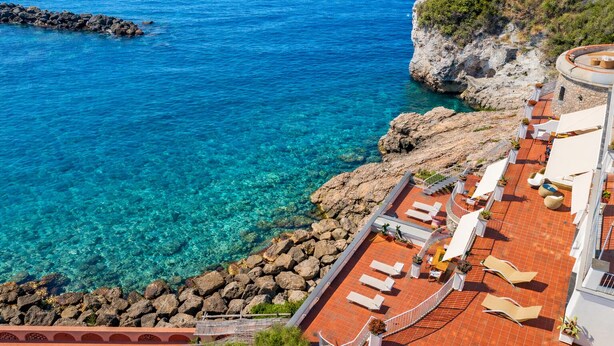 A coastal hotel with a terrace overlooking clear blue waters and rocky terrain.