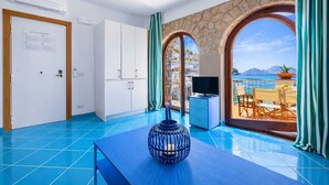 A room with a blue tiled floor, a television on a blue cabinet, a door with a notice, and a view of the sea through an archway.
