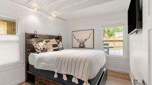 Une chambre avec une tête de lit en bois, un grand lit recouvert d’une couverture à rayures, un téléviseur fixé au mur et une fenêtre donnant sur une clôture en bois.