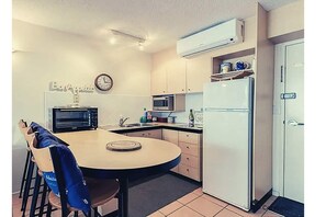 Compact kitchen with a round table and chairs, microwave, refrigerator, and clock.