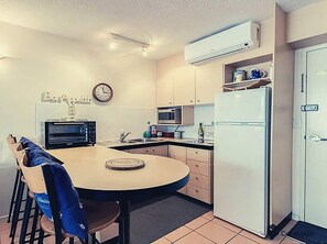 Compact kitchen with a round table and chairs, microwave, refrigerator, and clock.