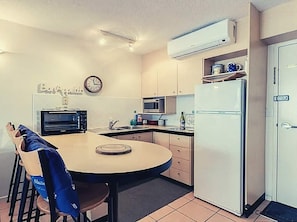 Compact kitchen with a round table and chairs, microwave, refrigerator, and clock.