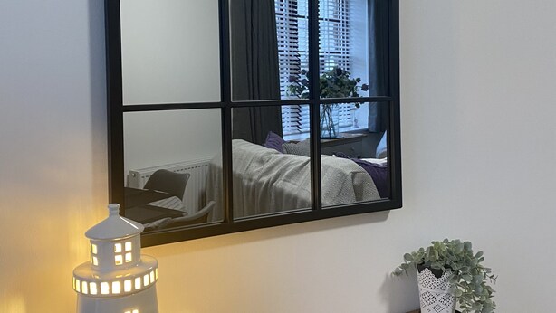 A room with a large mirror reflecting a bed and window, a small table with a book and a decorative lamp, and a potted plant. - The Howff - Lovely 2-bed Apartment in Anstruther (Anstruther)