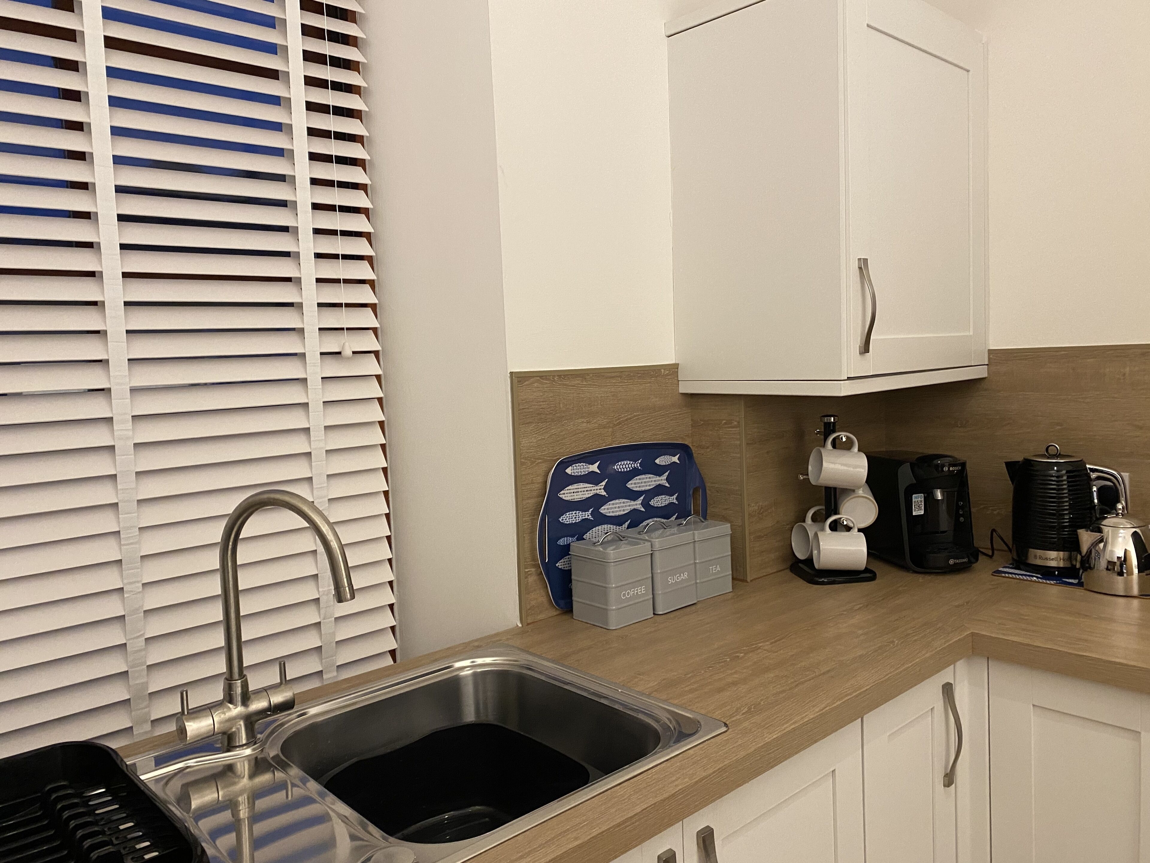 Een keuken met een roestvrijstalen spoelbak, witte kastjes, een koffiezetapparaat en een waterkoker.