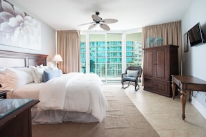 Habitación de hotel con una cama grande, ventilador de techo, vista de un edificio a través del ventanal, armario de madera y televisor en la pared.