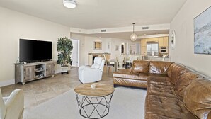A spacious living room with a large flat-screen TV, leather sofas, a white armchair, and a dining area in the background.