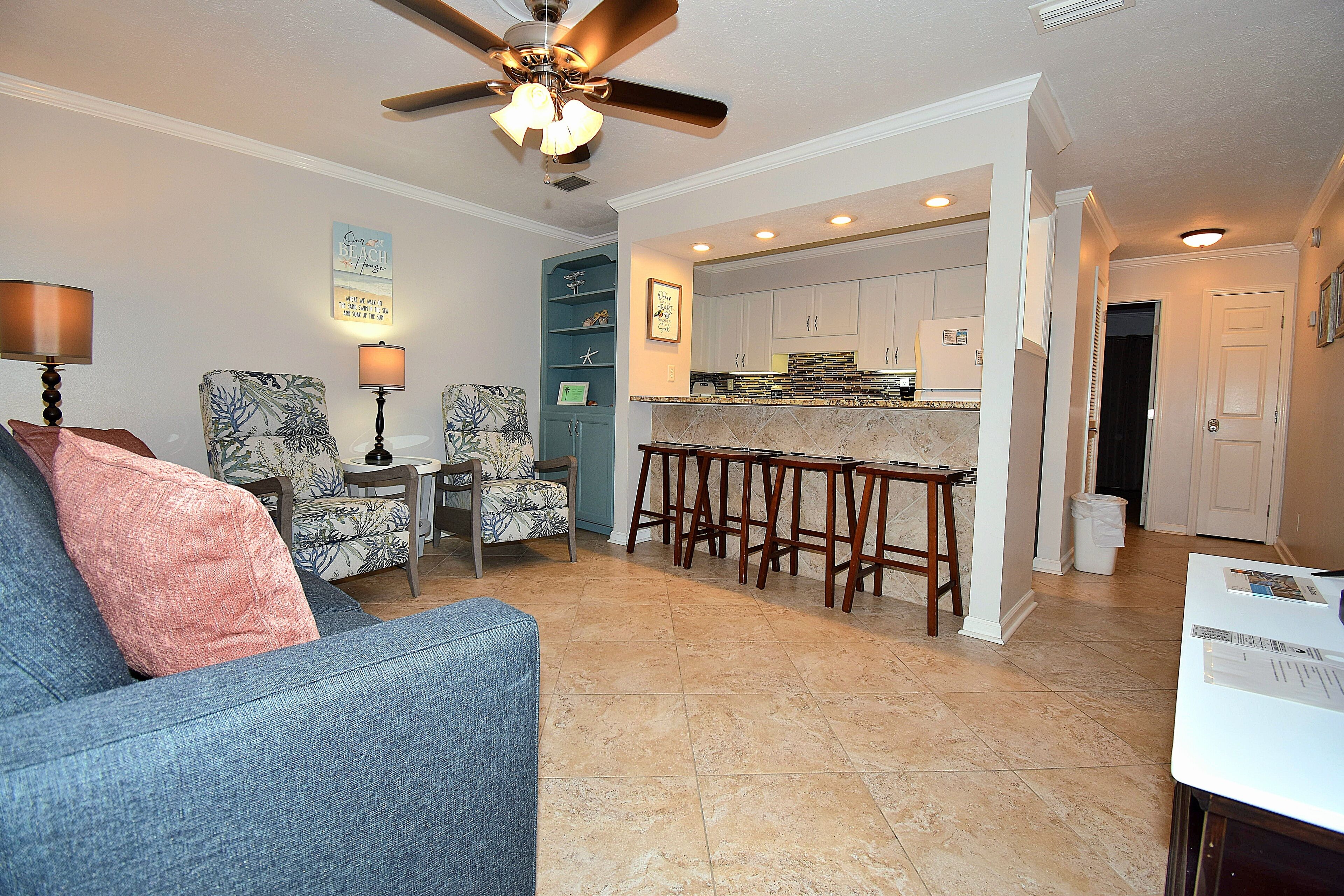 A living room with a sofa, chairs, a ceiling fan, and a kitchen area with a bar.