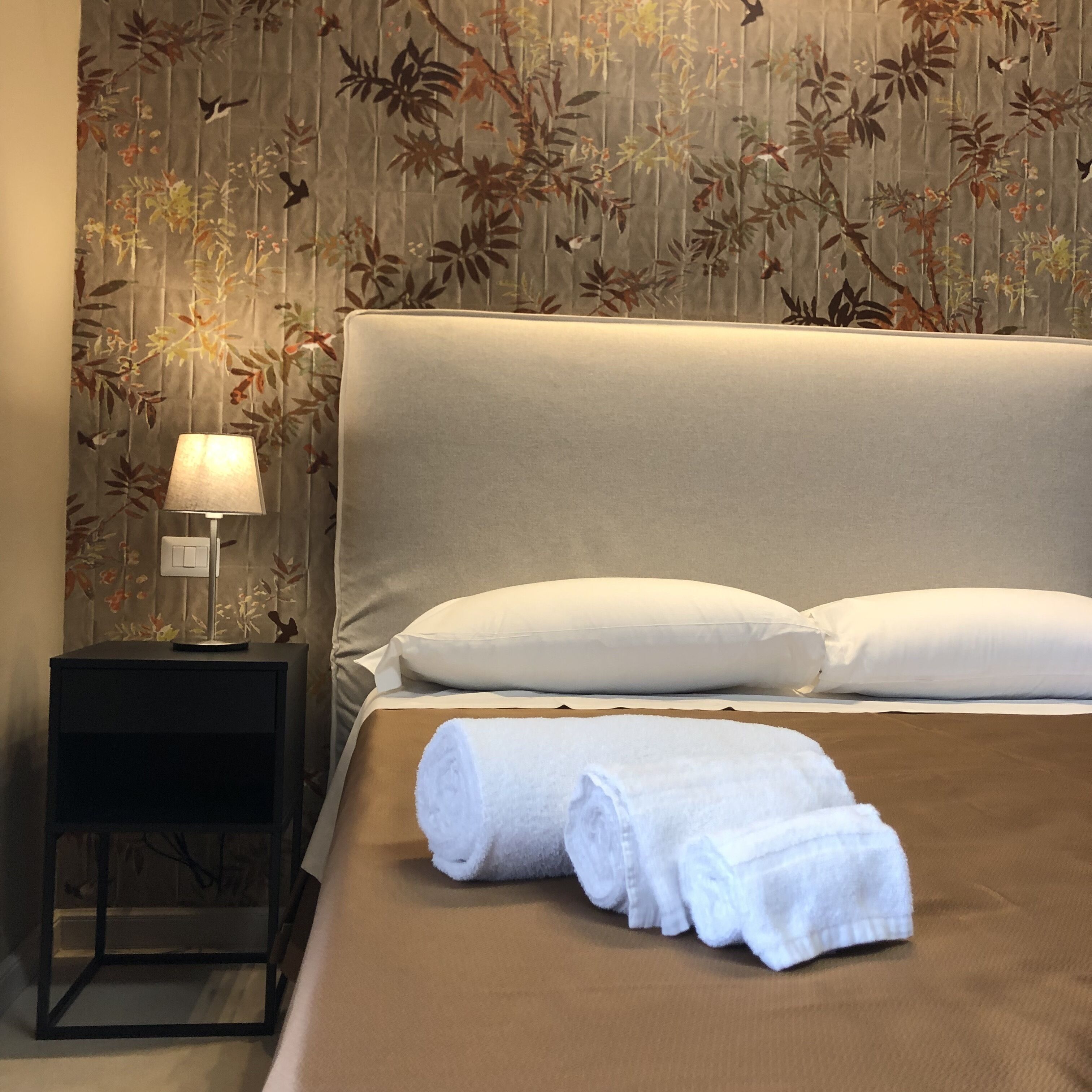 Een net opgemaakt bed met witte kussens en een opgerolde witte handdoek, een nachtkastje met een lamp, en bloemenbehang.