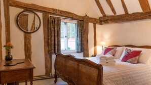 Ein Schlafzimmer mit Holz-Kopfteil, einem Nachttisch mit einer Vase, einem Fenster mit gemusterten Vorhängen und einem Bett mit weißen Leinentüchern und dekorativen Kissen.