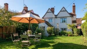 Eine Terrasse mit Tisch und Stühlen unter einem großen Sonnenschirm, umgeben von einem gepflegten Rasen und einem Haus mit mehreren Schornsteinen.