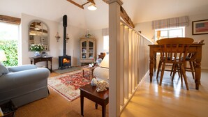 A cozy living room with a fireplace, a dining area with a wooden table and chairs, and a large window with a view of greenery. - The Stables -  A rural, romantic retreat in a particularly pretty countryside setting close to Castl (Great Maplestead)