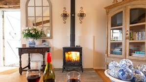 A cozy room with a wood-burning stove, a wooden cabinet with glass doors, and a vase with flowers on a table. - The Stables -  A rural, romantic retreat in a particularly pretty countryside setting close to Castl (Great Maplestead)