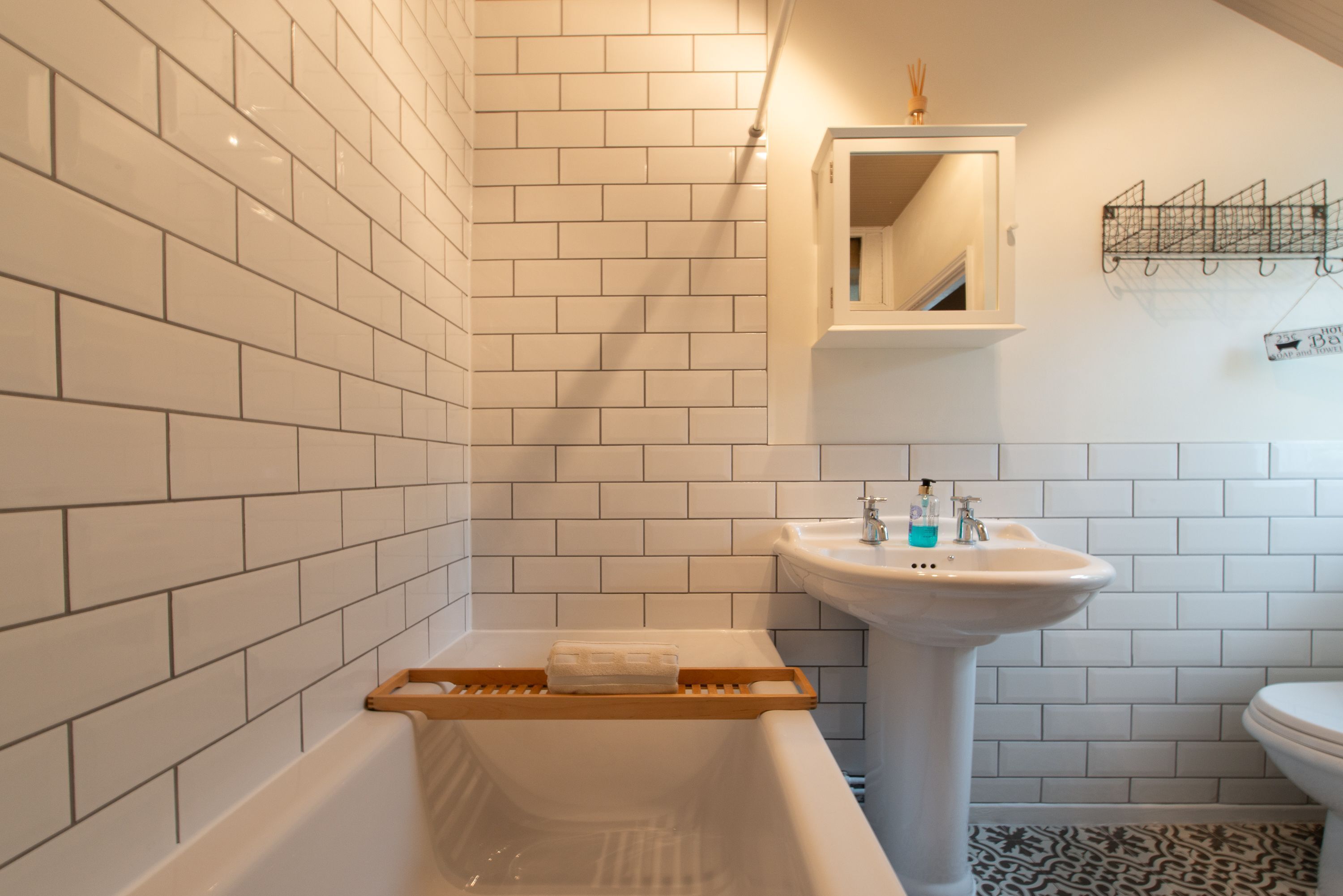 Une salle de bain avec des murs carrelés de blanc, un lavabo sur colonne, un plateau de bain en bois et une petite étagère contenant des articles de toilette.