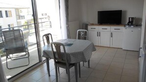 Et lille, lyst værelse med balkon, spisebord med stole, fjernsyn og tekøkken.