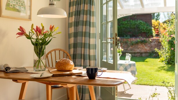 A wooden dining table with a loaf of bread, a vase of flowers, and a mug. - A beautiful little Victorian House in one of Suffolk’s prettiest villages. (Cavendish)