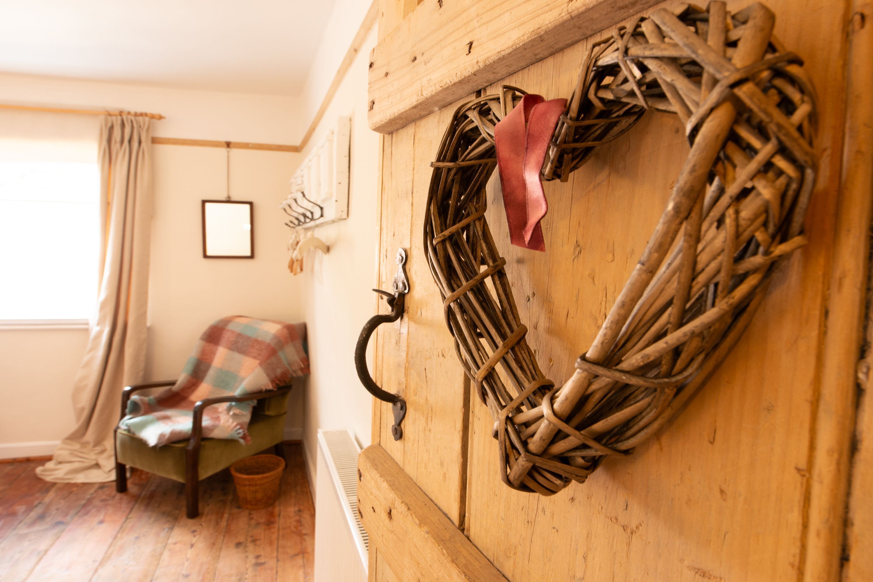 A room with a wicker wreath on a wooden wall, a plaid chair, and a window with curtains.