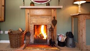 A fireplace with a fire, a wooden mantel, a mirror above it, and a lamp to the right. - A beautiful little Victorian House in one of Suffolk’s prettiest villages. (Cavendish)