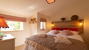 A bedroom with a large bed, a bedside table, a window with curtains, and a wooden headboard with wall-mounted lights. - A beautiful little Victorian House in one of Suffolk’s prettiest villages. (Cavendish)
