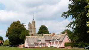 A pink building with a thatched roof and a clock tower in the background. - A beautiful little Victorian House in one of Suffolk’s prettiest villages. (Cavendish)