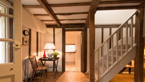 Una escalera de madera con barandal blanco conduce a un piso superior. Un sillón de madera y una mesa con una lámpara y una planta están ubicados cerca de una puerta en el piso inferior.