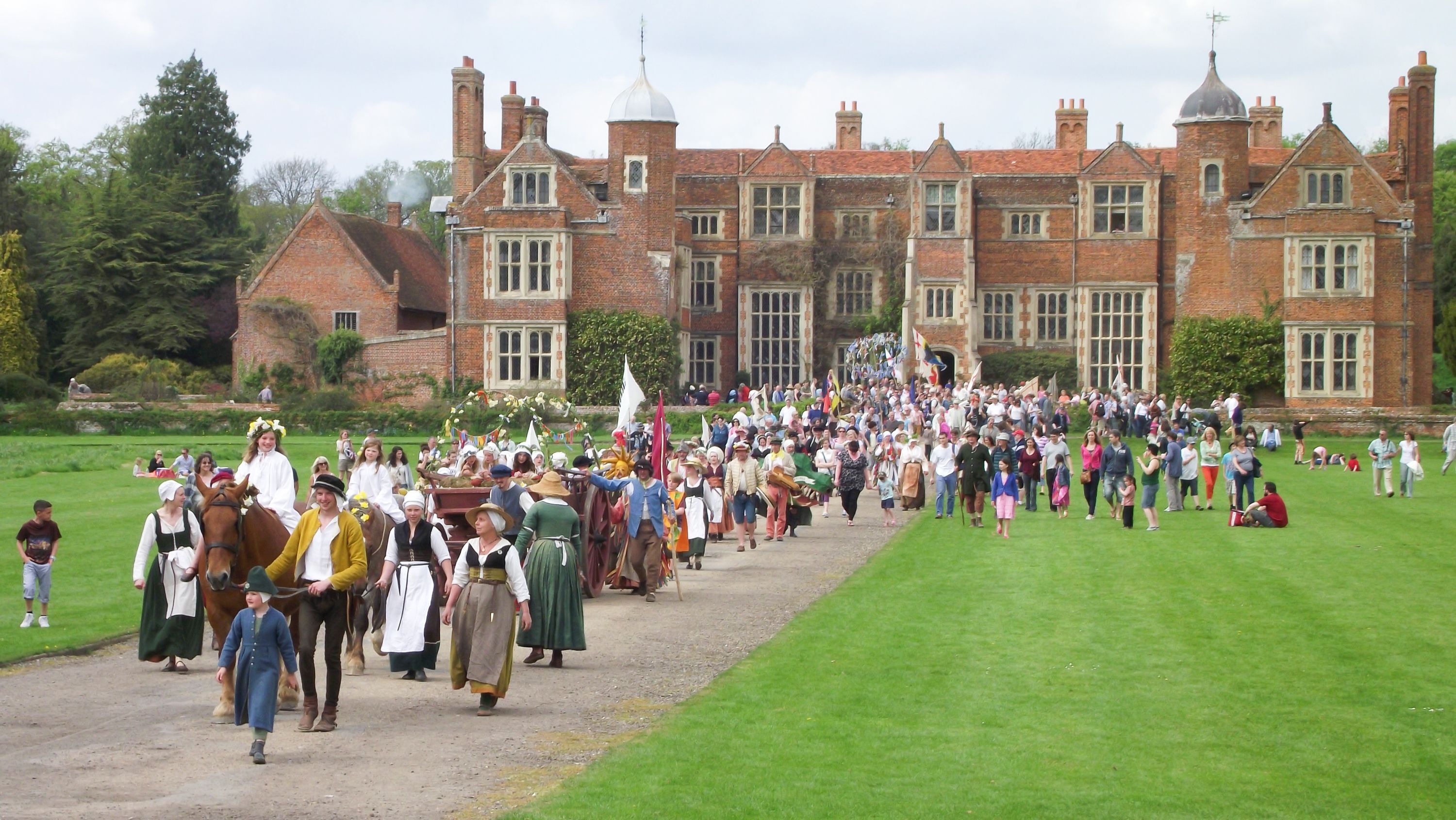 A large group of people in historical costumes walking in front of a large, traditional brick building with multiple windows and a prominent tower.
