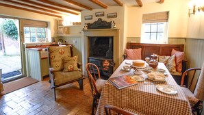 A cozy dining area with a checkered tablecloth, wooden chairs, a fireplace, and a view of the garden through a window.