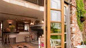 A cozy kitchen with a brick fireplace, a dining area with white metal chairs, and a large window overlooking a garden. - Dons Barn - A romantic holiday cottage with beautiful, beamed interiors. Wow. (Edwardstone)
