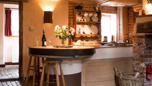 A kitchen with a brick wall, wooden stools, a wine bottle, and a vase of flowers. - Dons Barn - A romantic holiday cottage with beautiful, beamed interiors. Wow. (Edwardstone)