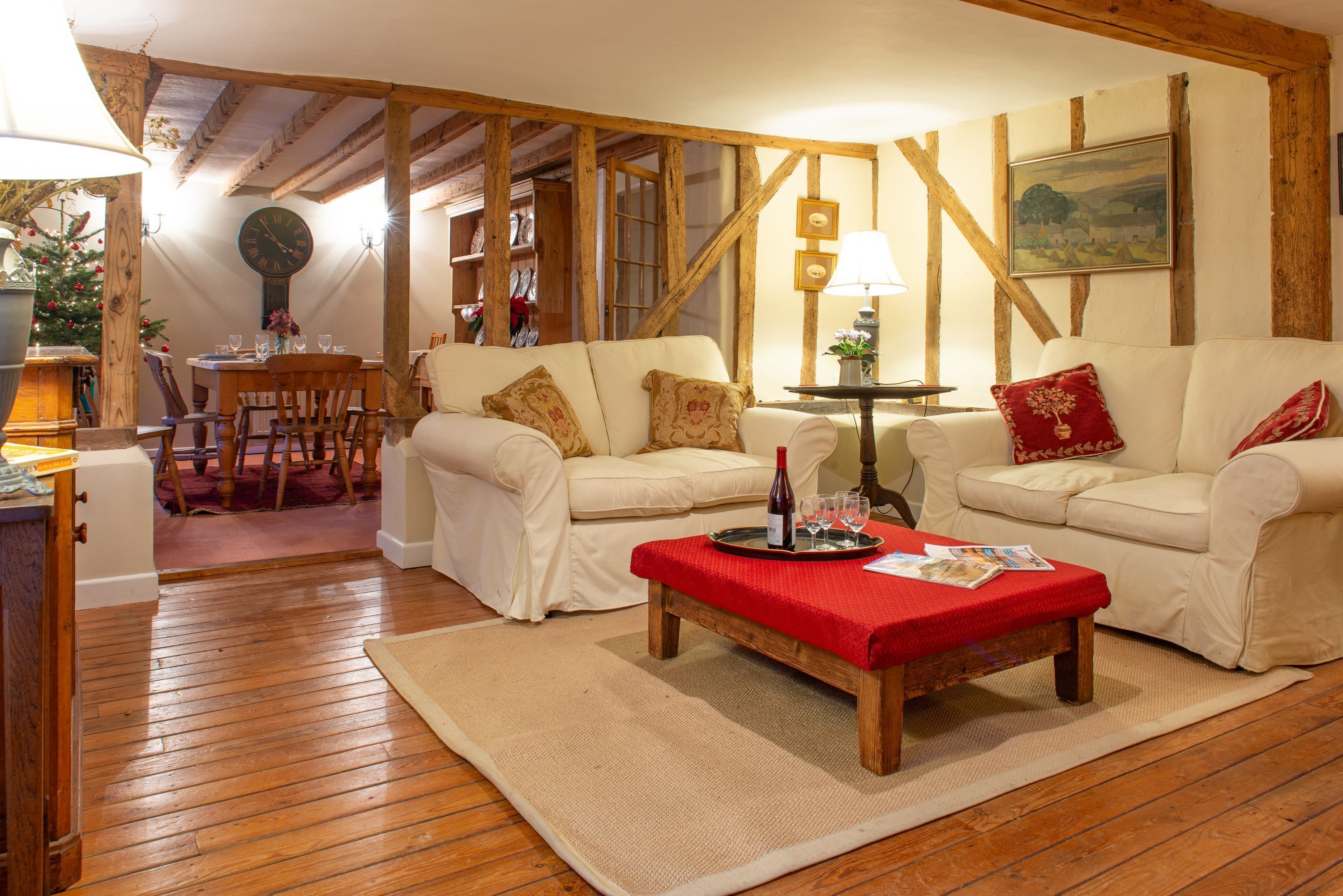 Un salón acogedor con vigas de madera, dos sofás color crema, una mesa de centro de madera con un mantel rojo y una zona de comedor con mesa y sillas de madera.