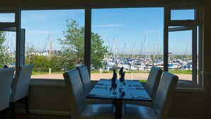 A dining area with a table set for four, overlooking a marina with numerous boats. - Remote, Comfort, Sea Views, and the romance of the beautiful Essex Marshes - The Wobbin (Tollesbury)