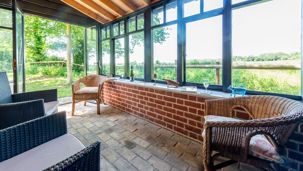 A covered patio area with wicker furniture, a brick wall, and large windows offering a view of the outdoors. - Remote, Comfort, Sea Views, and the romance of the beautiful Essex Marshes - The Wobbin (Tollesbury)