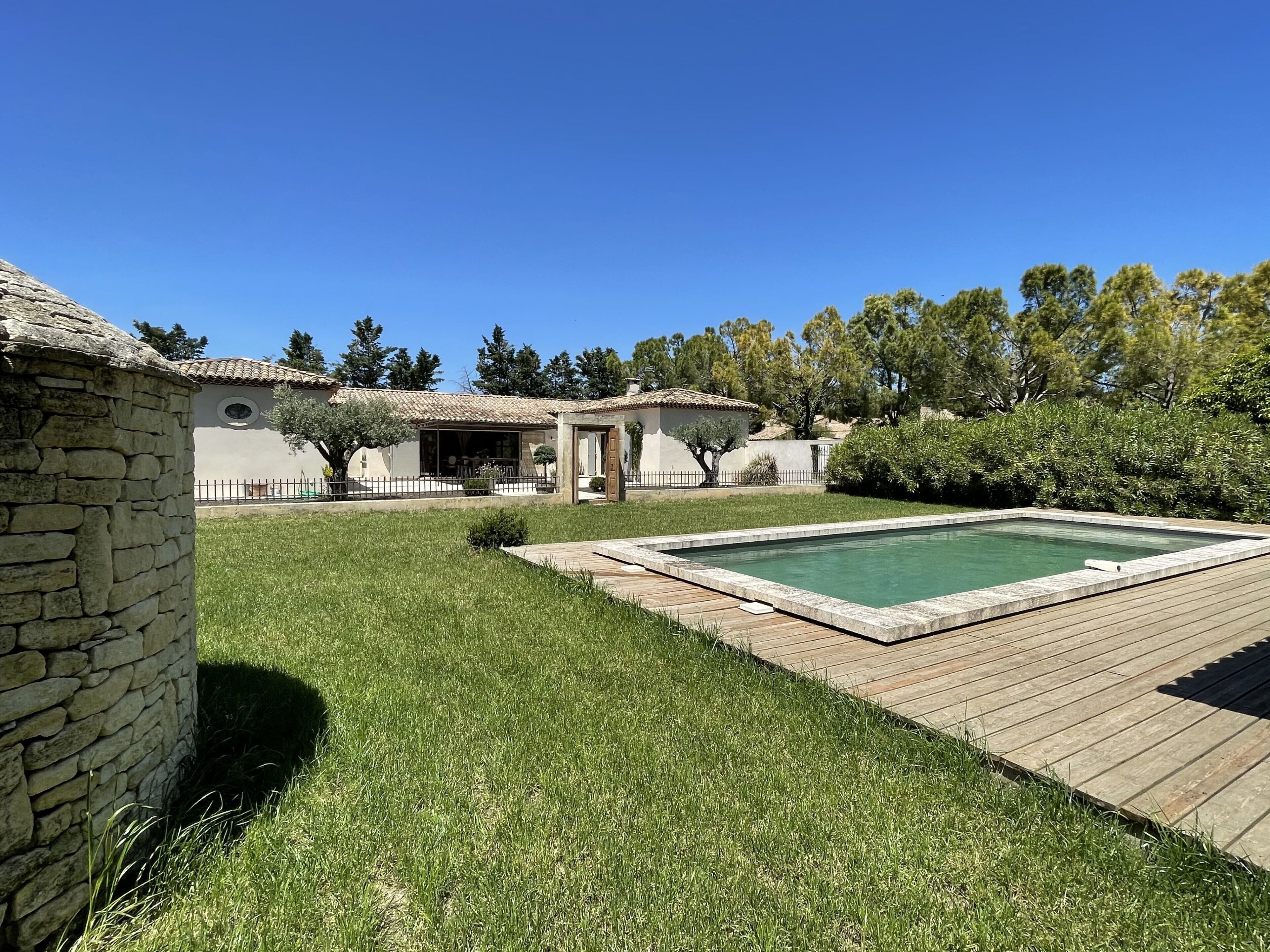 Un bâtiment en pierre avec un toit en tuiles, une piscine dotée d’une terrasse en bois et une pelouse avec des arbres en arrière-plan.